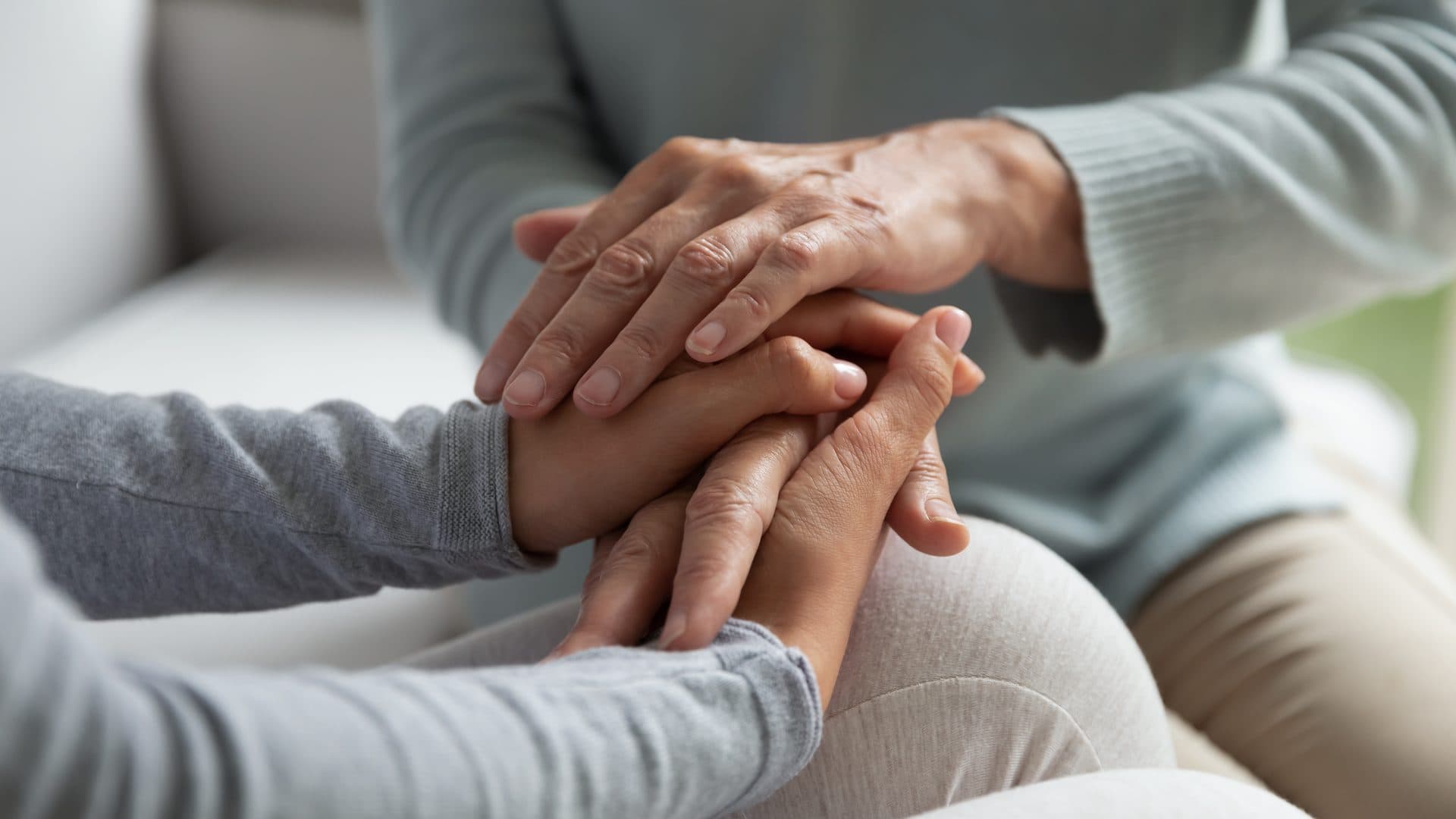 Nahaufnahme: Die Hände einer älteren Person umschließen die Hand einer jüngeren, beide sitzen in hellen Strickpullovern nebeneinander im weichen Tageslicht