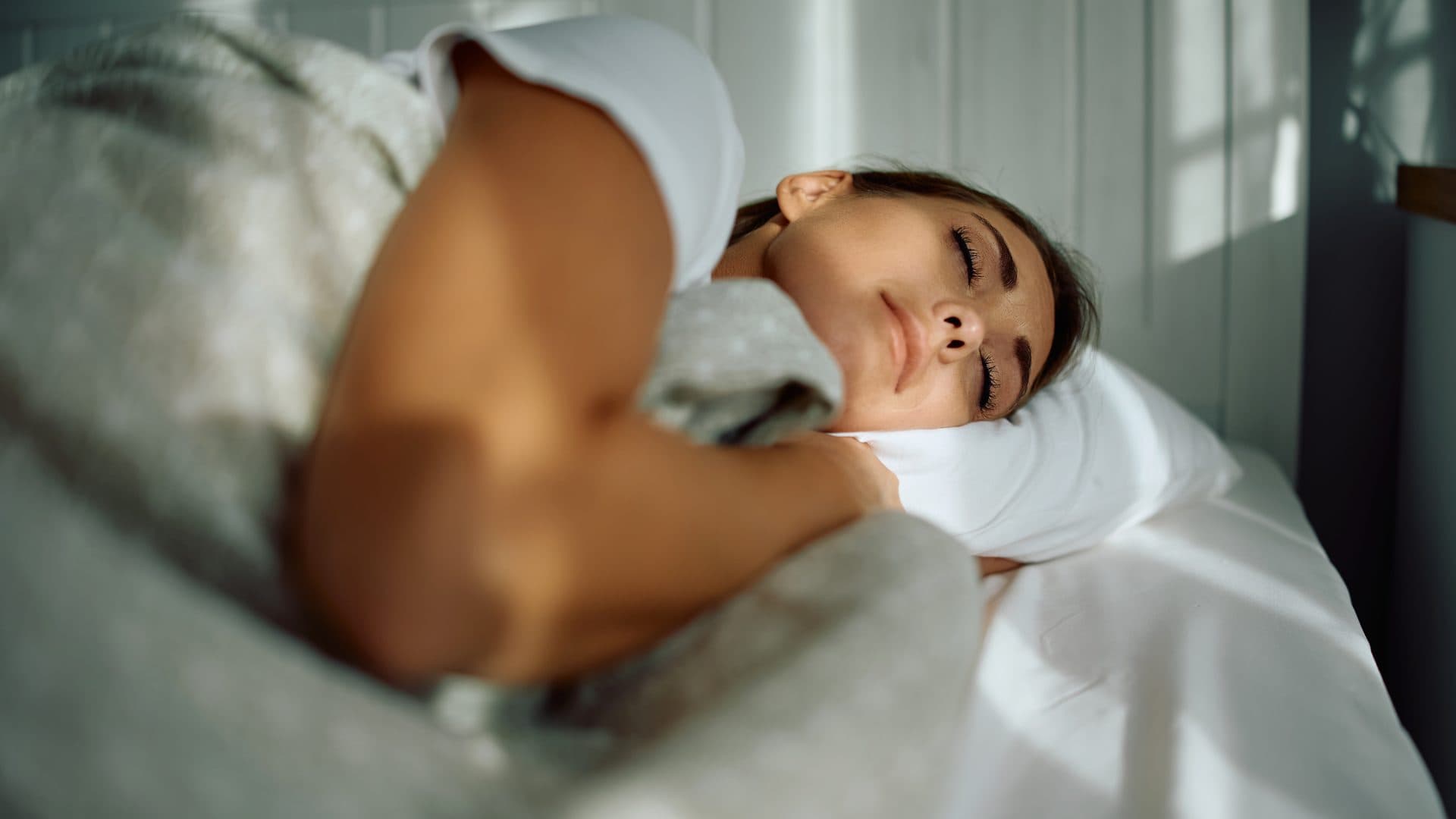 Frau schläft auf der Seite im Bett, den Kopf auf einem weissen Kissen, einen Arm über der hellen Bettdecke, weiches Tageslicht im Schlafzimmer