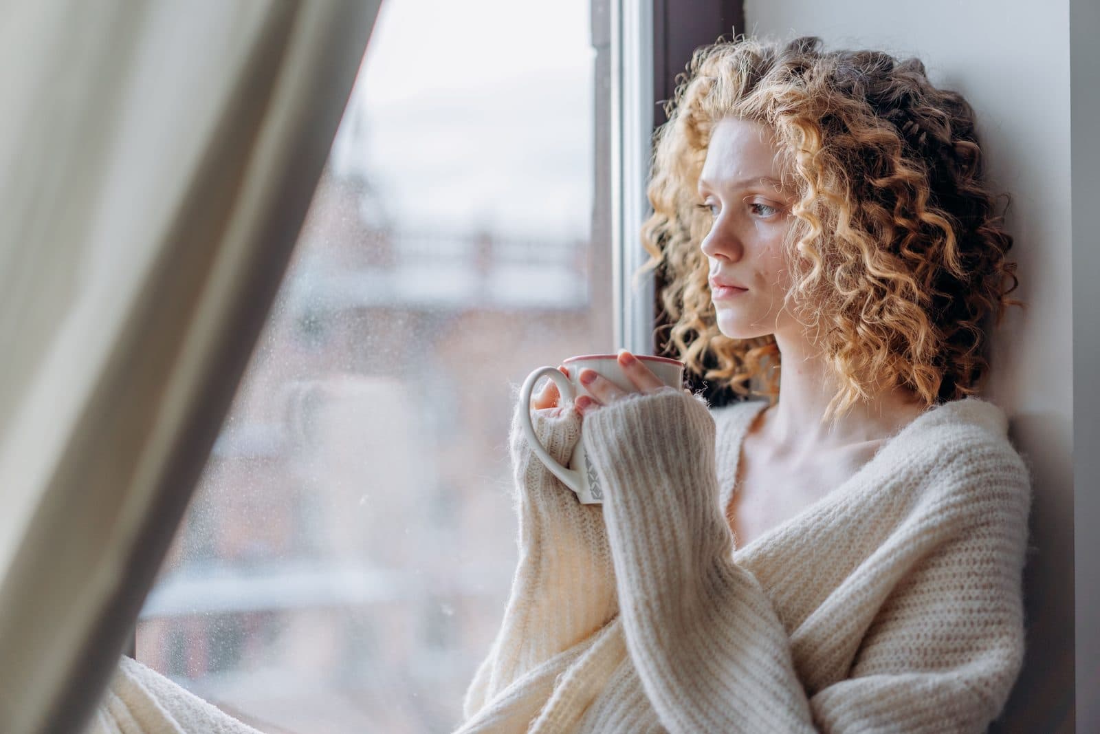 Junge Frau mit blonden Locken sitzt in einer Fensternische und hält mit beiden Händen eine weiße Tasse; die Ärmel ihres Strickpullovers sind über die Finger gezogen