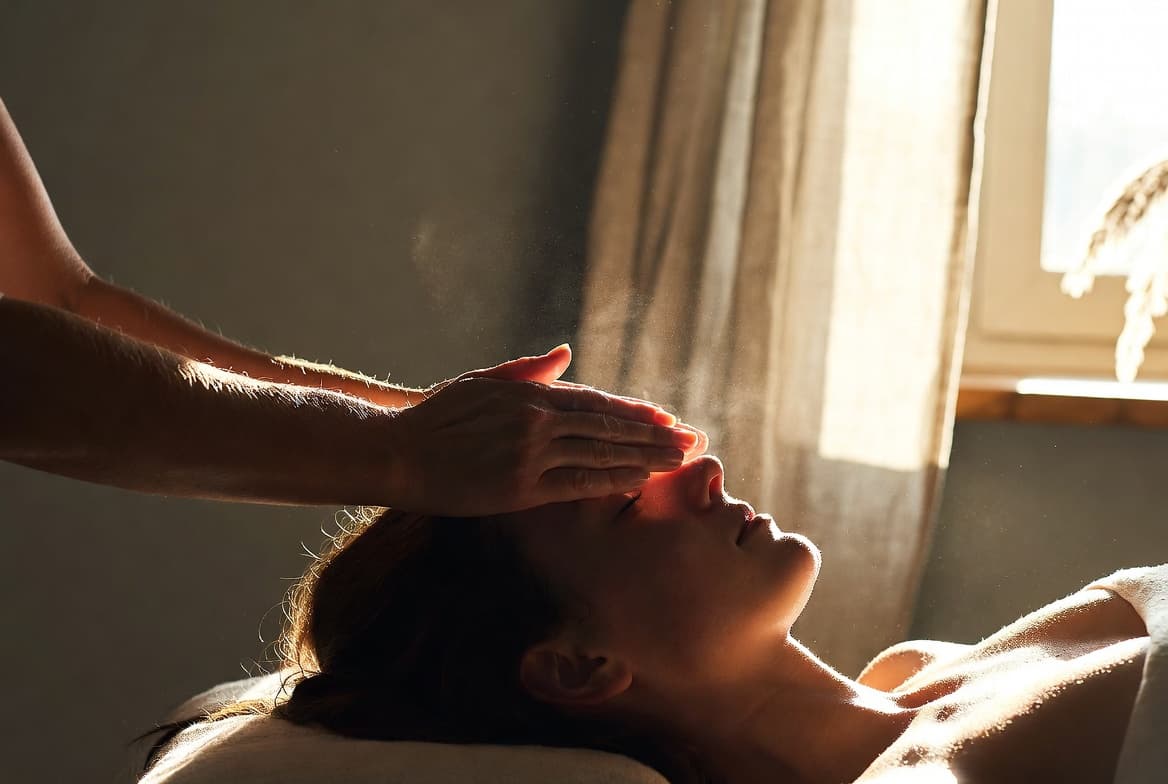 Spokojna sesja reiki — dłonie praktyka uniesione nad leżącą osobą, miękkie naturalne światło, neutralne wnętrze