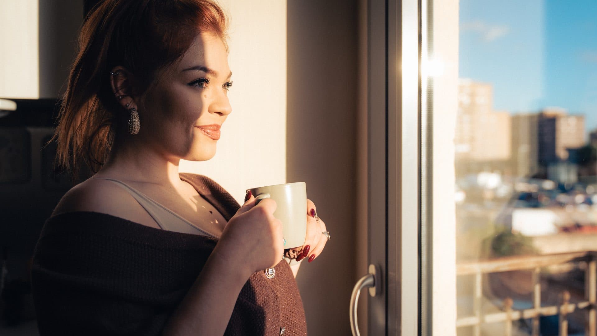 Junge Frau mit rötlichem Haar steht im warmen Gegenlicht am Fenster, hält eine helle Tasse mit beiden Händen und blickt nach draußen