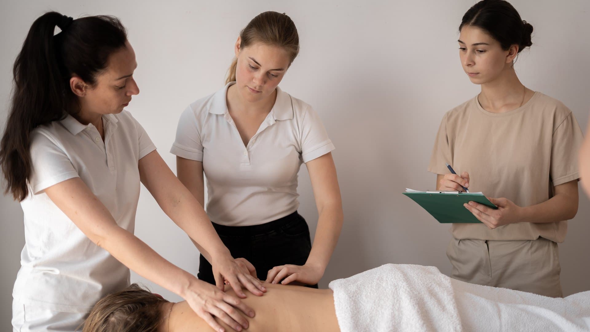 Drei Fachpersonen in heller Berufskleidung an einer Massageliege: eine führt die Hände einer Lernenden auf dem Rücken einer liegenden Person, eine dritte notiert auf einem Klemmbrett