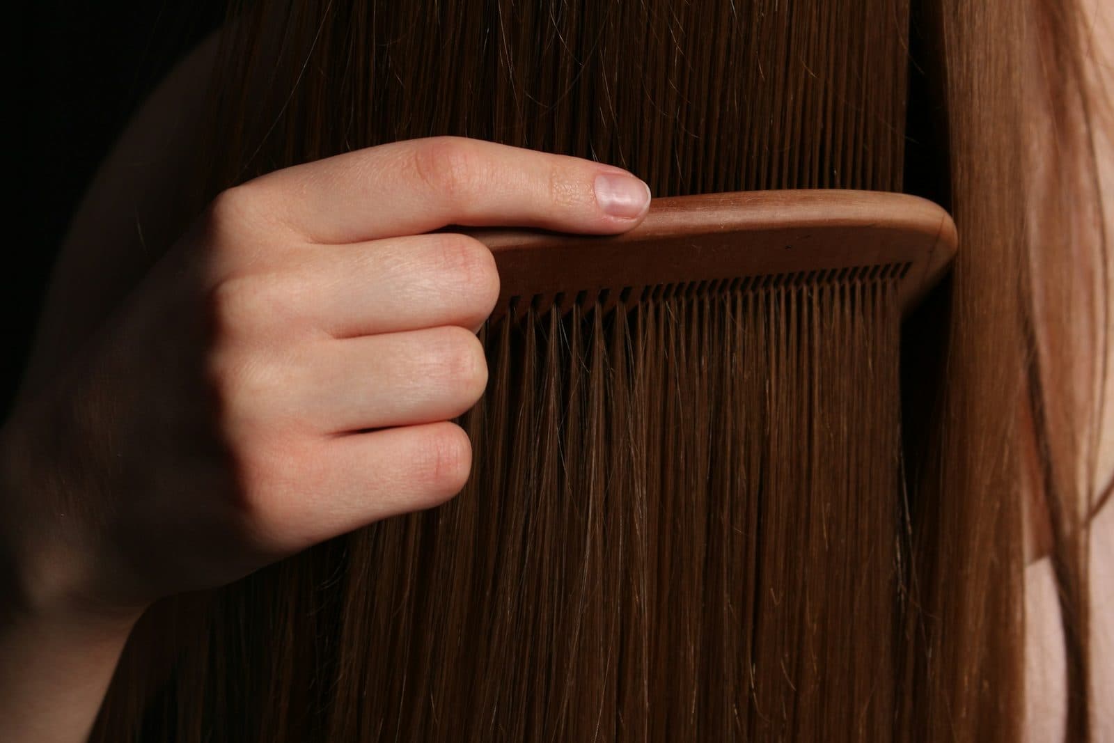 Eine Hand zieht einen hellen Holzkamm langsam durch langes, glattes braunes Haar; der Hintergrund bleibt dunkel, nur die einzelnen Strähnen leuchten auf