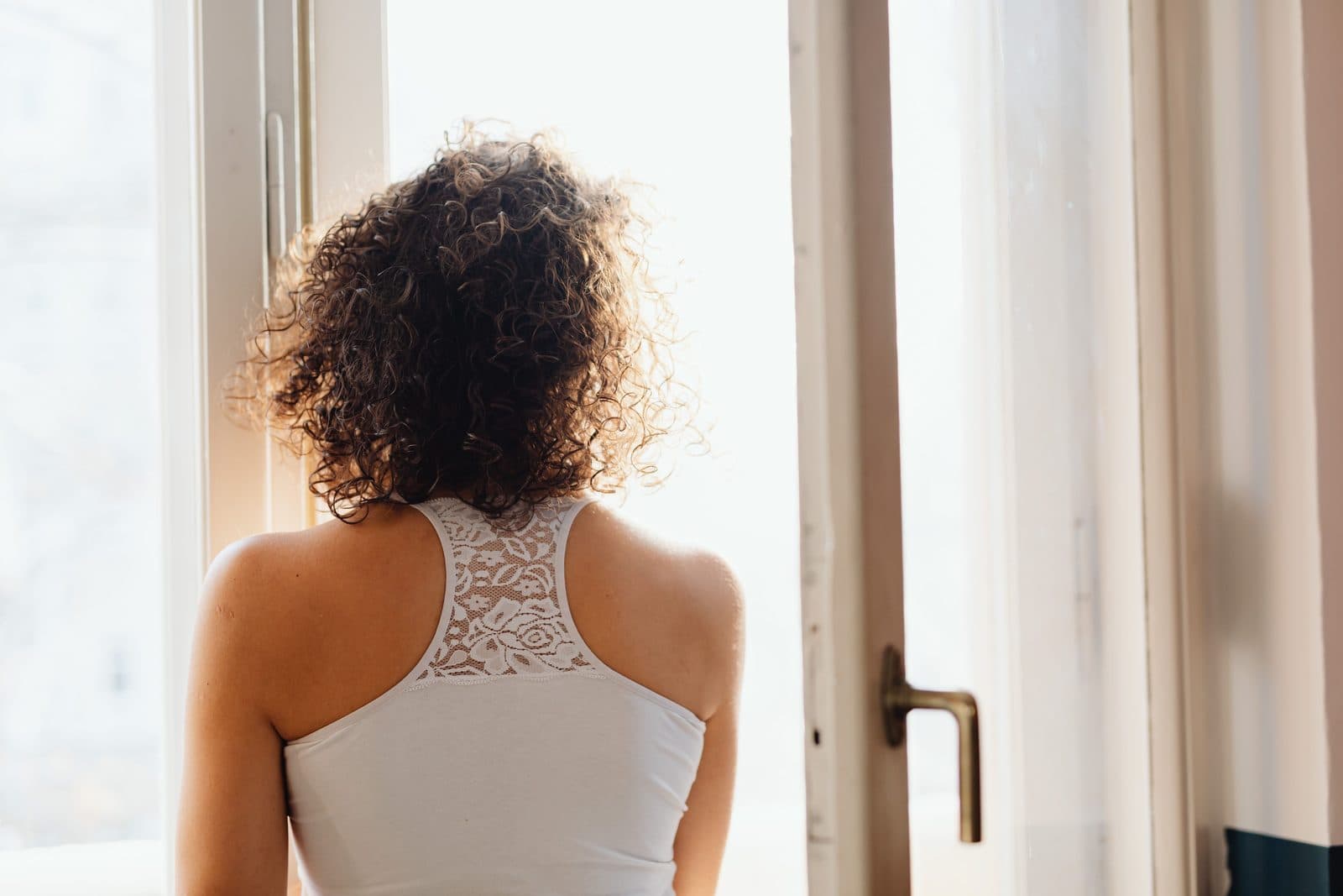 Eine Frau mit lockigem Haar steht von hinten gesehen in einem weißen ärmellosen Top an einem offenen Fenster; helles Tageslicht fällt über ihre Schultern in den Raum