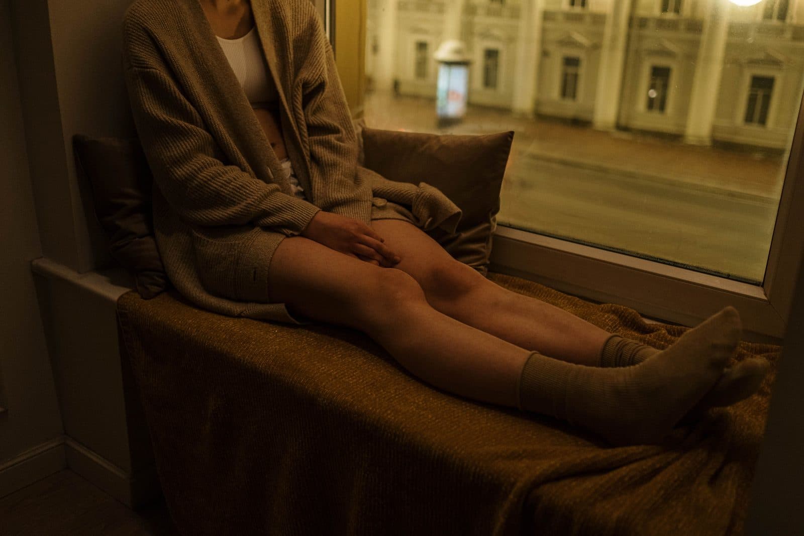 Person in Strickjacke und Socken sitzt mit ausgestreckten Beinen auf einer gepolsterten Fensterbank, eine Hand ruht auf dem Knie; draußen warmes Abendlicht und eine beleuchtete Hausfassade