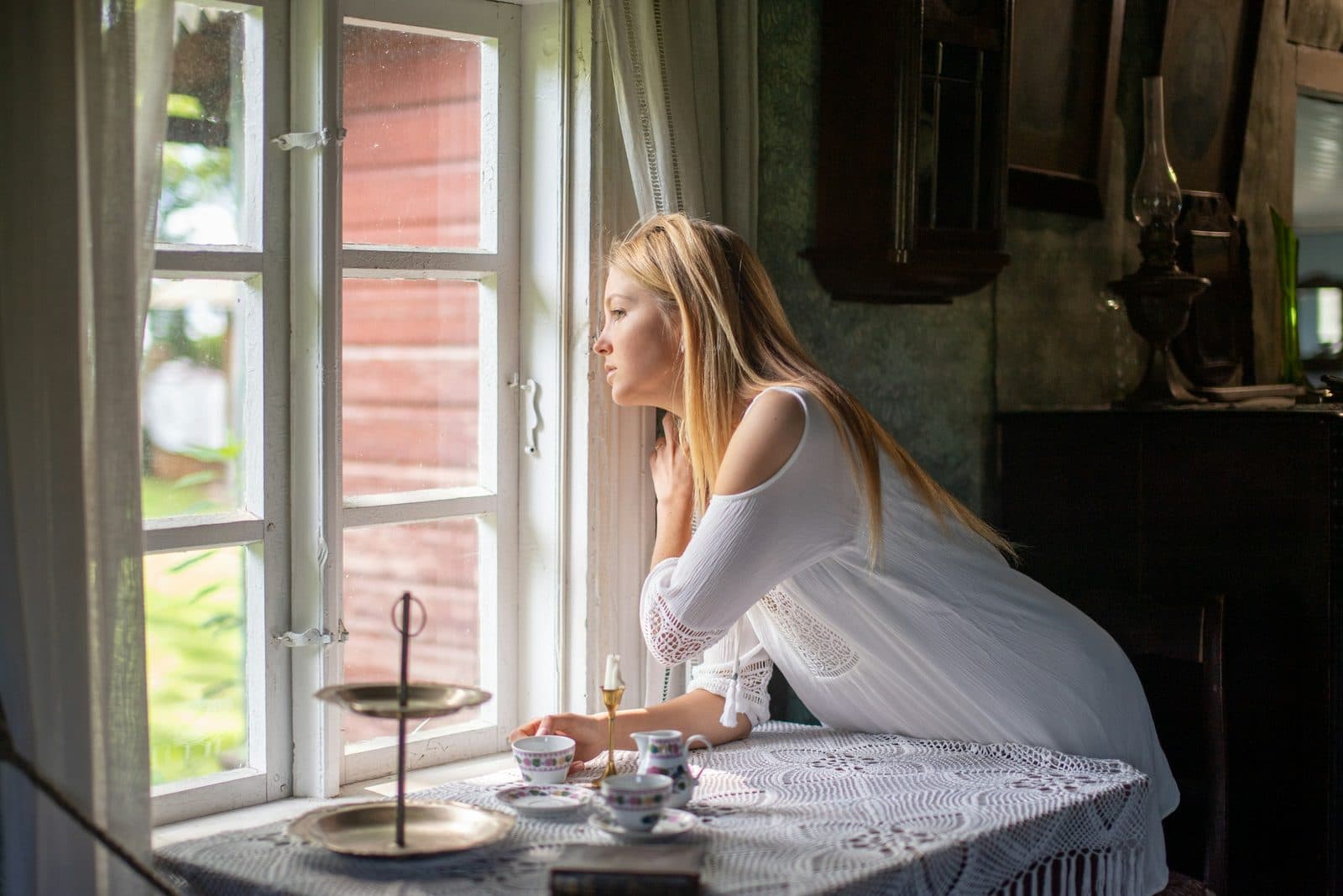 Eine Frau im weißen Kleid stützt sich an einem Tisch am offenen Fenster eines Landhauses auf und blickt hinaus; auf der Spitzendecke stehen kleine Porzellantassen